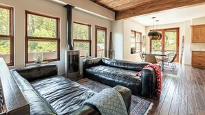 Een ruime woonkamer met een leren bank, een eethoek met tafel en stoelen, grote ramen met uitzicht op bomen, en een houten plafond.