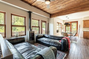 Un ampio soggiorno con divano in pelle, zona pranzo con tavolo e sedie, ampie finestre con vista sugli alberi e soffitto rivestito in legno.