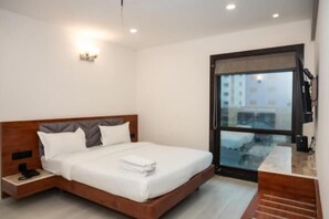 A modern hotel room with a wooden bed, a bedside table with a phone, a large window with a view, and a flat-screen TV.