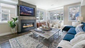 A living room with a fireplace, a television mounted above it, a sofa, and a coffee table. - Steps from Town Lift & Main Street! Luxe amenities! (Park City)