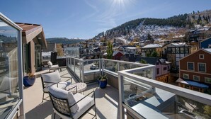 A balcony with outdoor seating, a glass railing, and a view of a town with buildings and a hill. - Steps from Town Lift & Main Street! Luxe amenities! (Park City)