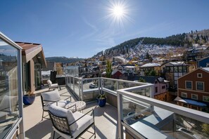 Ein Balkon mit Sitzgelegenheit im Freien, einer Glastrennwand und Blick auf eine Stadt mit Gebäuden und einem Hügel.