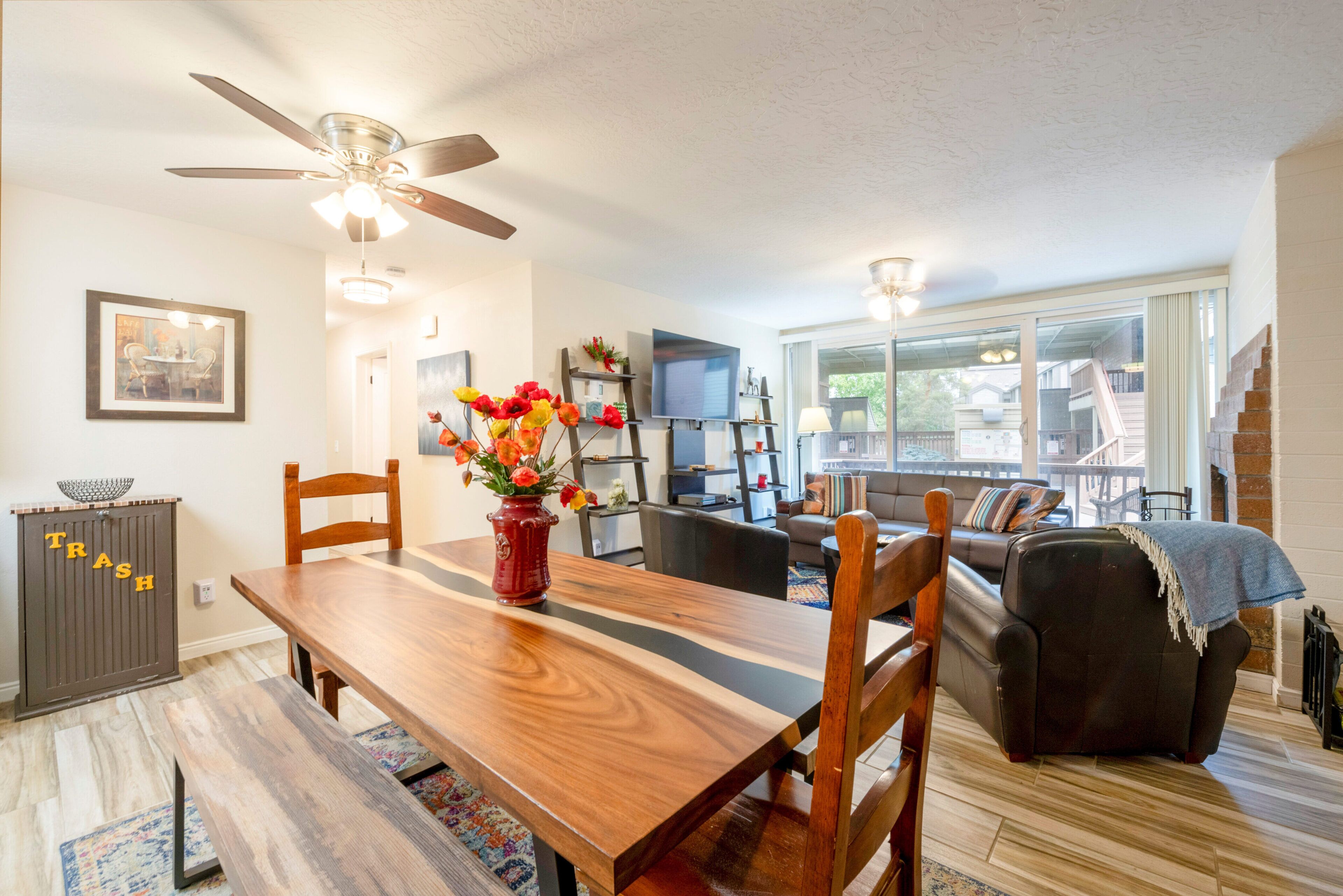 A spacious living room with a wooden dining table, a sofa, and a TV. There is a ceiling fan, a fireplace, and a large window.