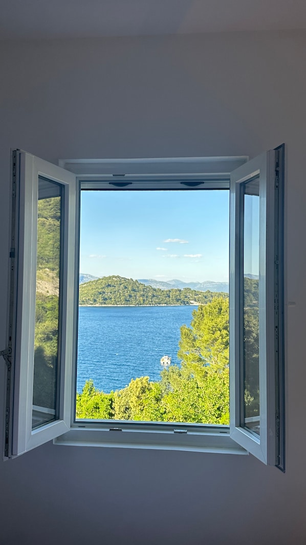 A window with a clear view of a body of water, greenery, and distant hills.