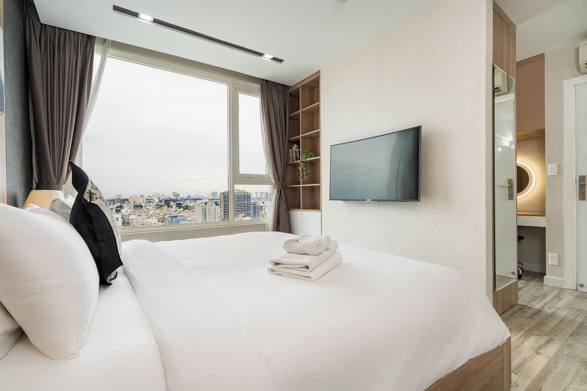 Ett hotellrum med en stor säng, en tv som är monterad på väggen, utsikt över staden och en bokhylla i trä.