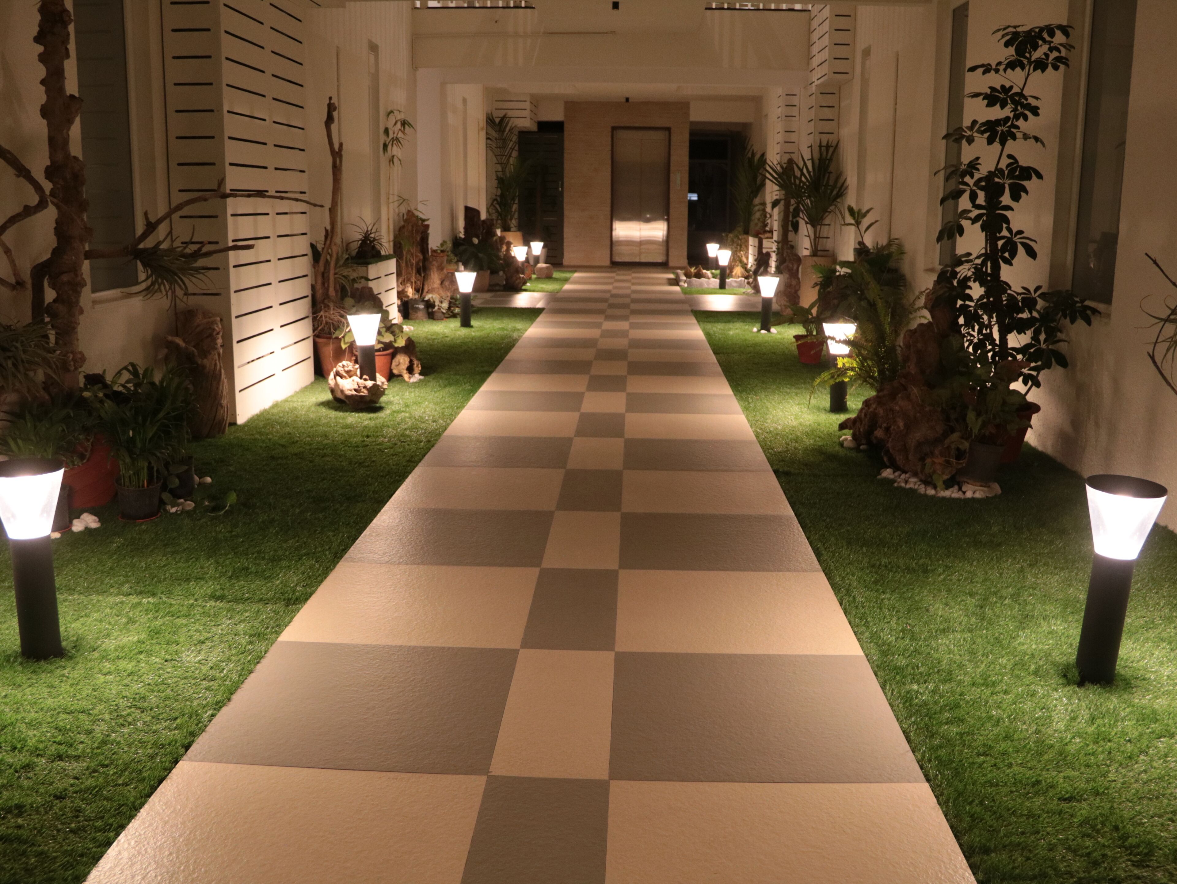 A hotel corridor with a checkered floor, potted plants, and wall-mounted lights.