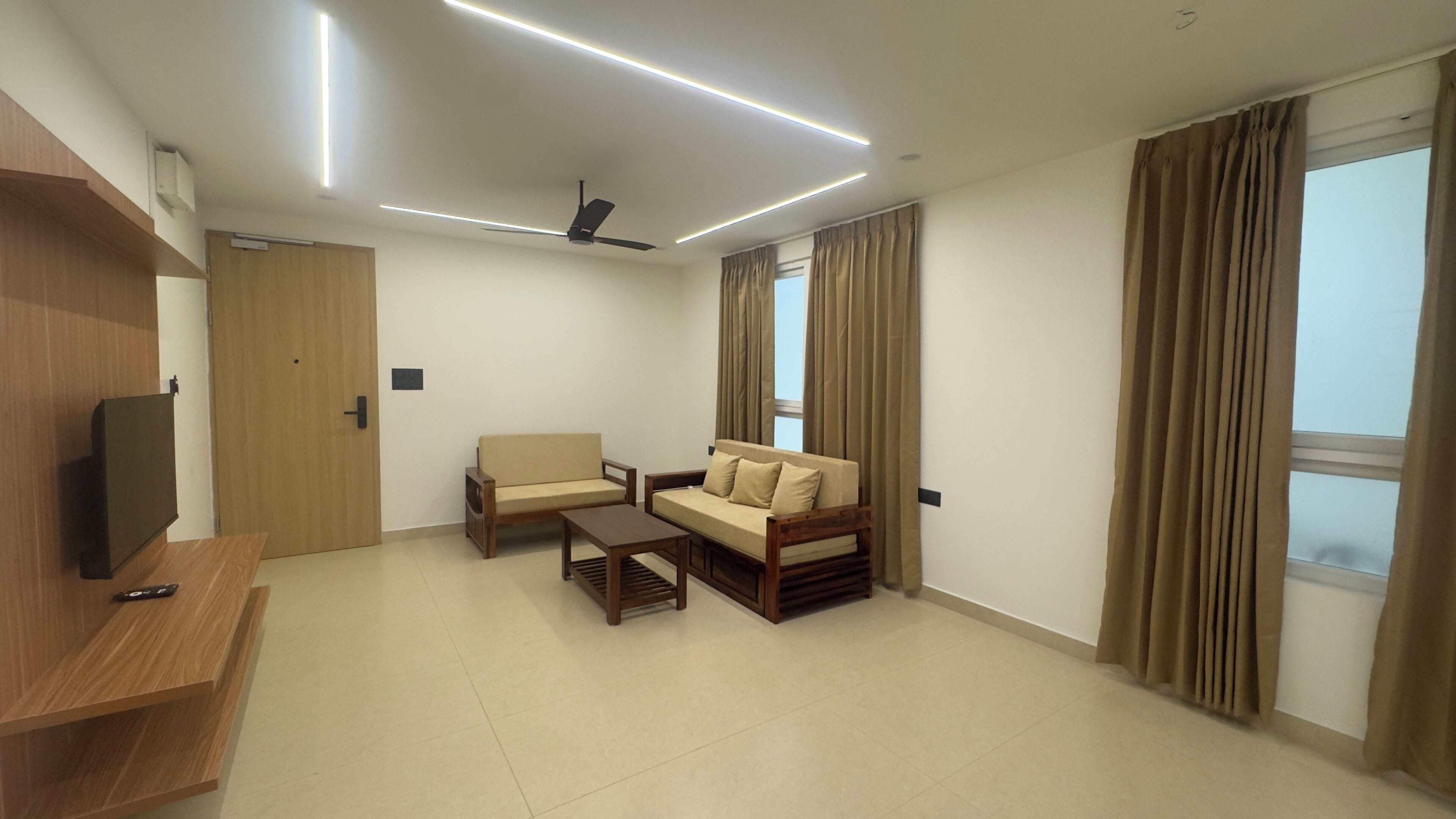 Una sala de estar moderna con sofá, sillones, una mesa de centro, televisor sobre un mueble de madera, ventilador de techo y ventanas amplias con cortinas.