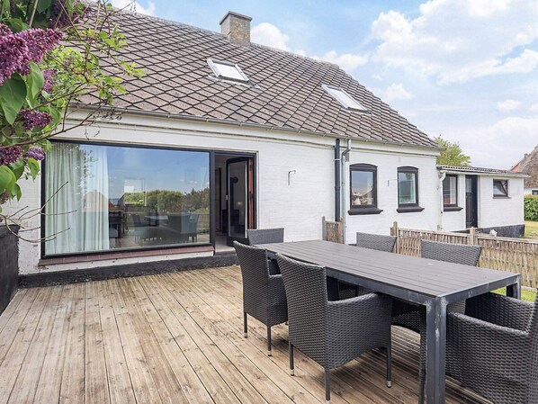 Une maison dotée d’une grande porte vitrée donnant sur une terrasse, d’une table à manger avec des chaises et d’une façade blanche.