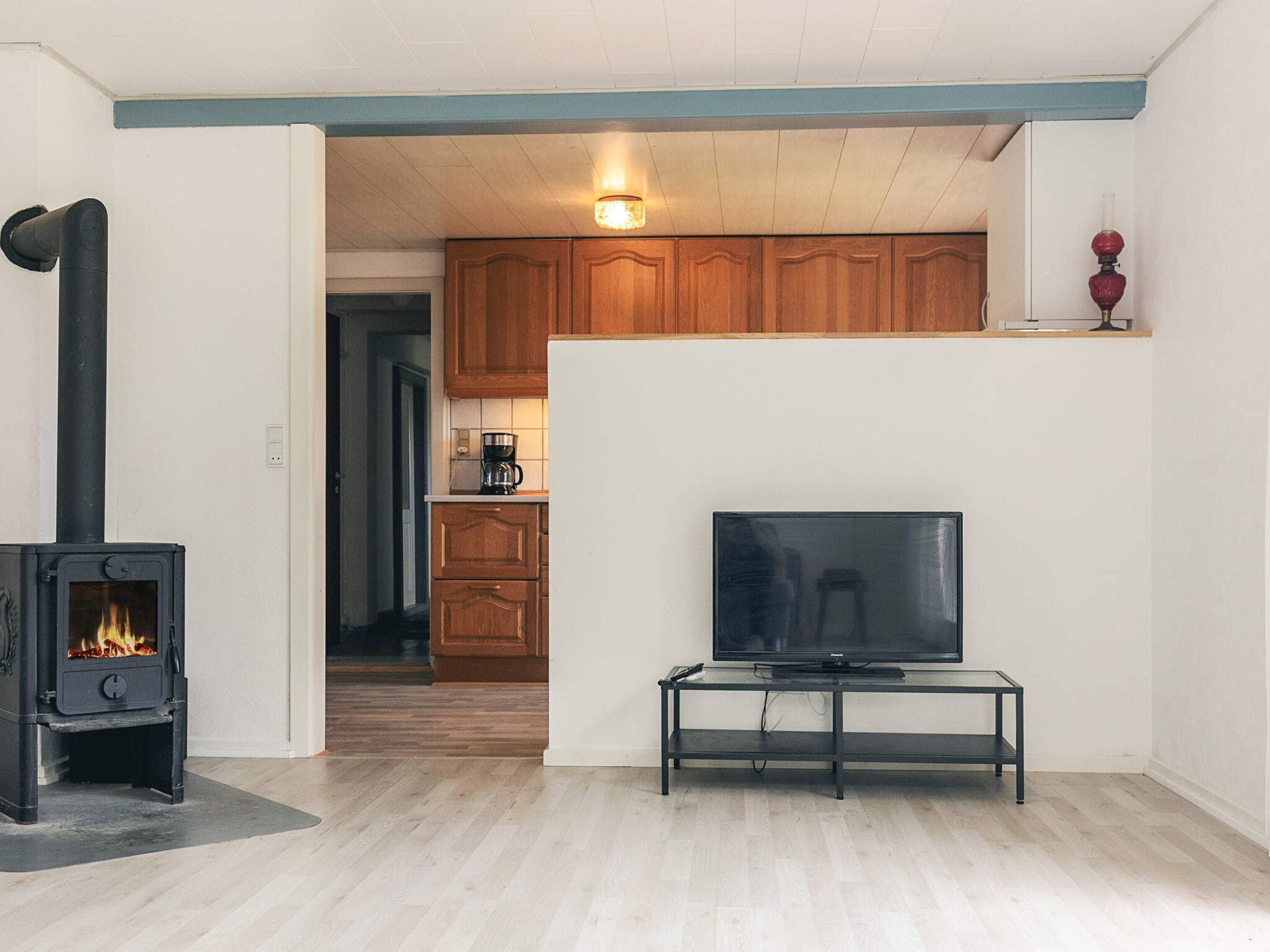 Una sala de estar con un estufa de leña negra, un televisor sobre un mueble bajo, y una cocina al fondo.