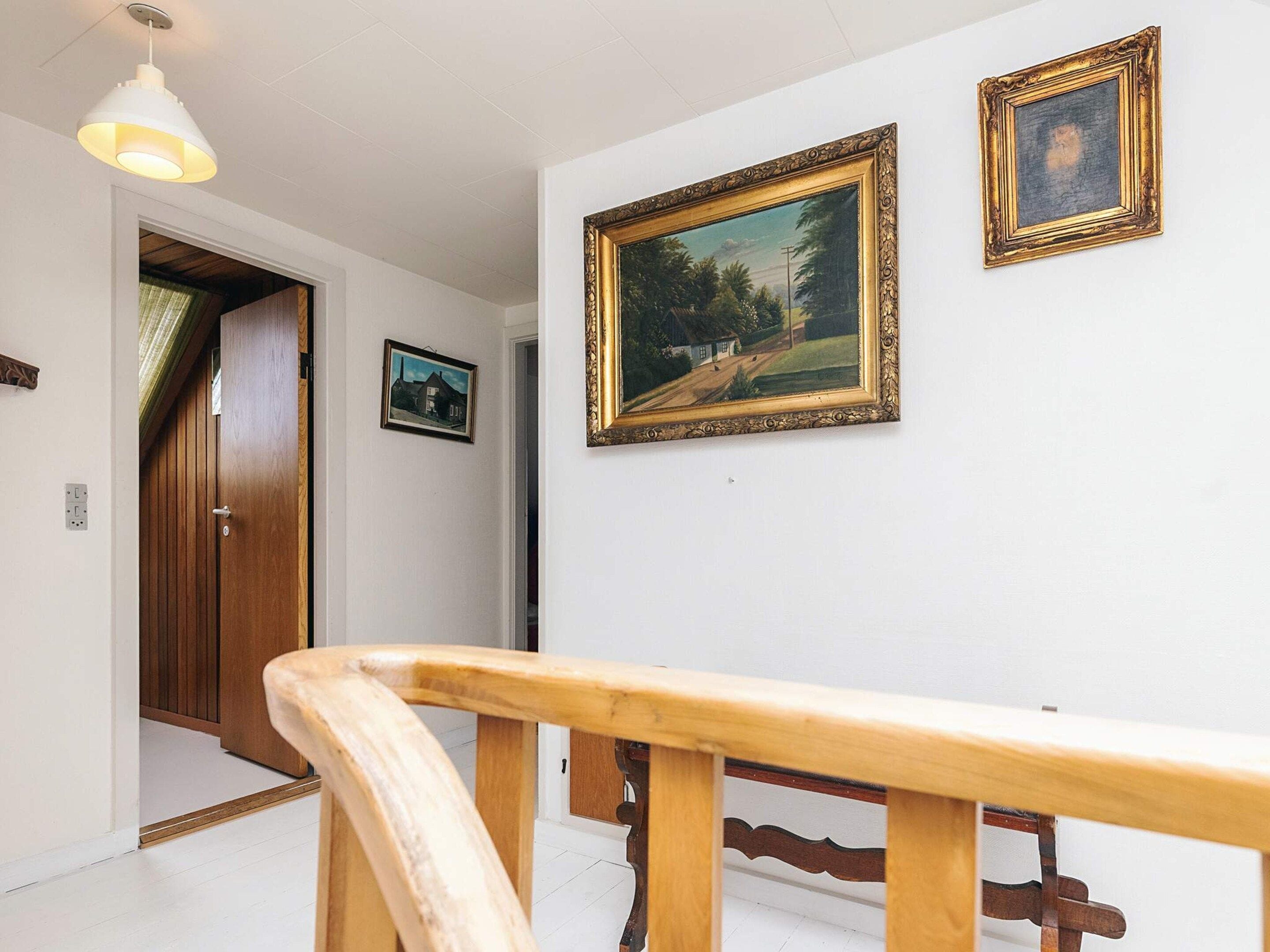 Un pasillo con puerta de madera, pared blanca y dos cuadros enmarcados, y barandilla de madera.