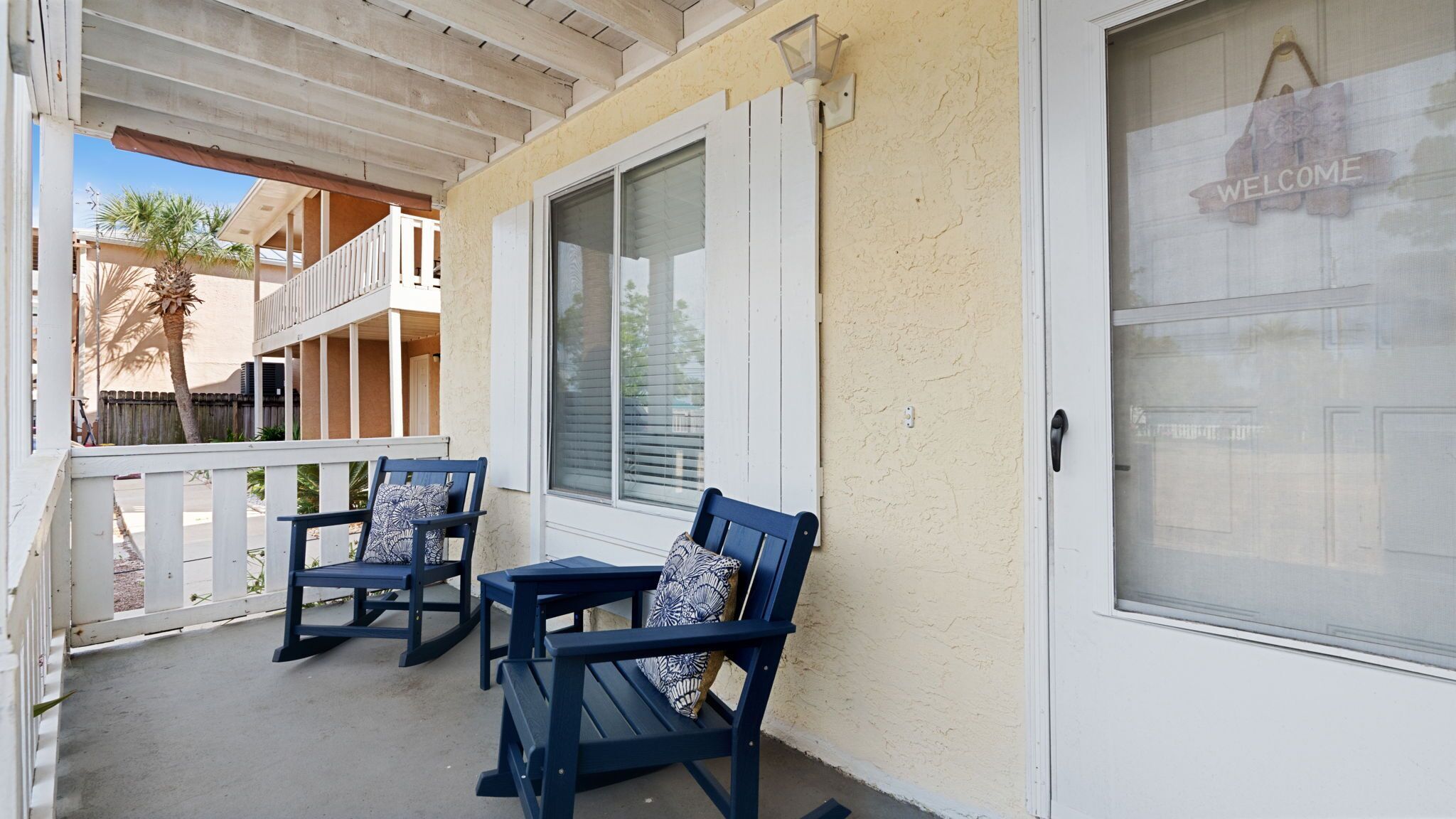 Eine überdachte Veranda mit zwei dunkelblauen Sesseln und einem Tisch, ein 'WELCOME'-Schild an der Tür und Blick auf ein Gebäude mit einem Balkon.