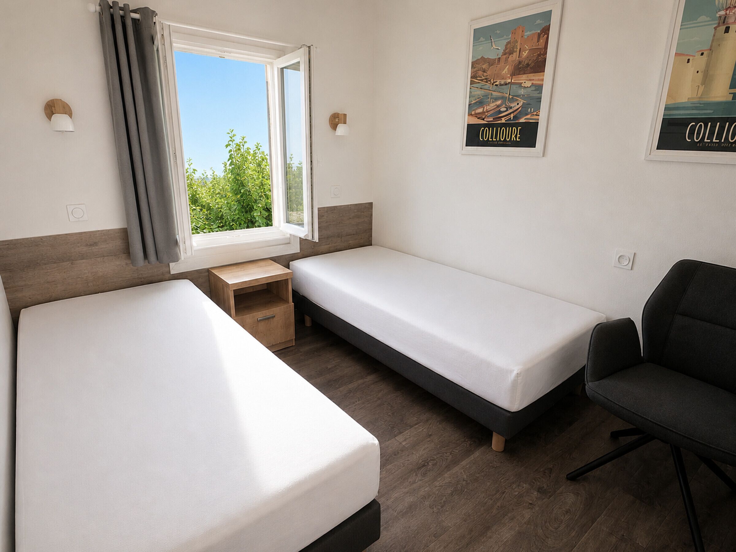 Una habitación con dos camas individuales, una mesita de noche de madera, un escritorio, una silla y una ventana con cortinas.