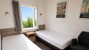 Een kamer met twee eenpersoonsbedden, een klein houten nachtkastje, een bureau, een stoel en een raam met gordijnen.