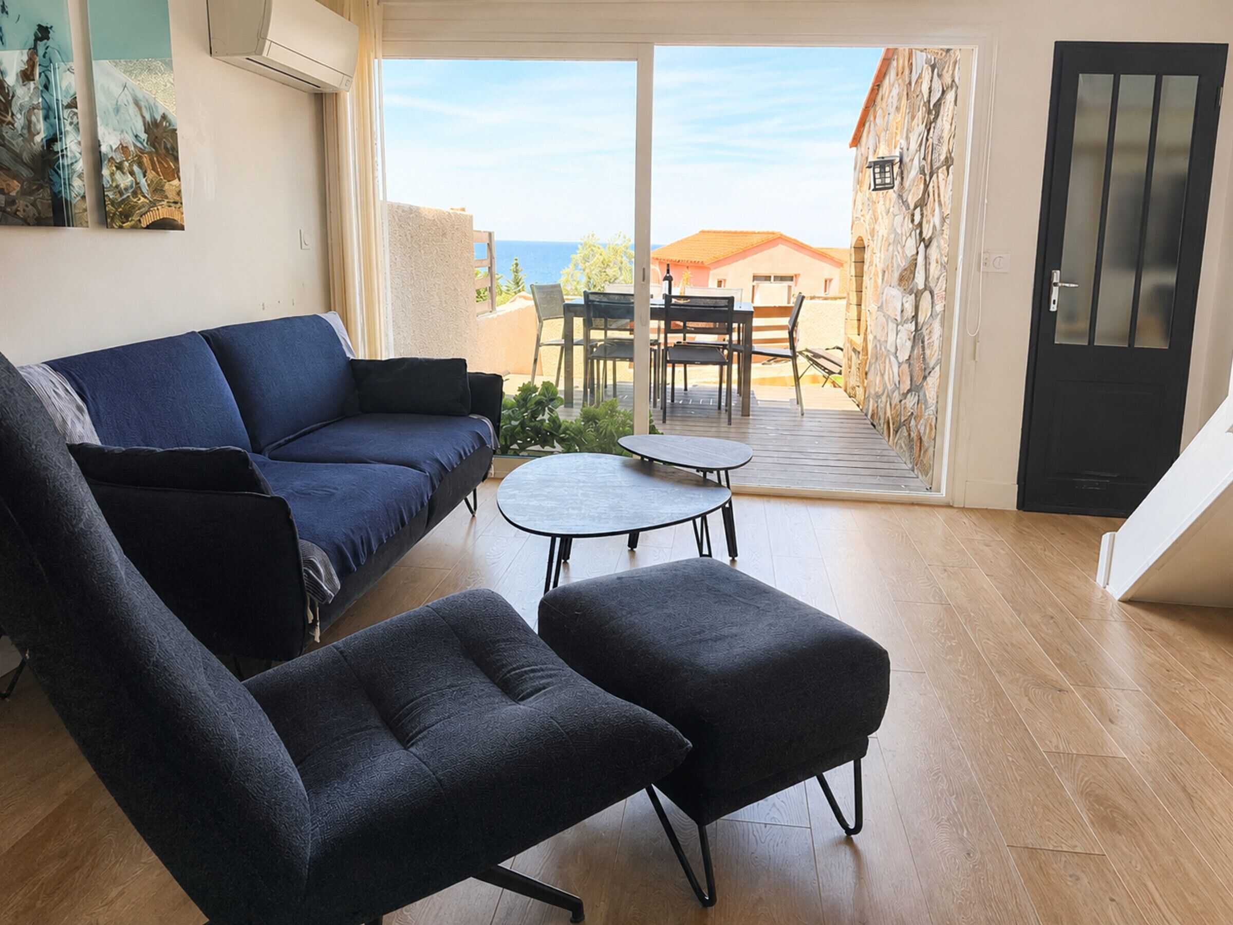 A living room with a sofa, two armchairs, a small table, and a view of the sea through a glass door.