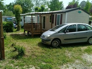 Una casa móvil con una pequeña terraza, un coche estacionado adelante y una antena parabólica en el techo.