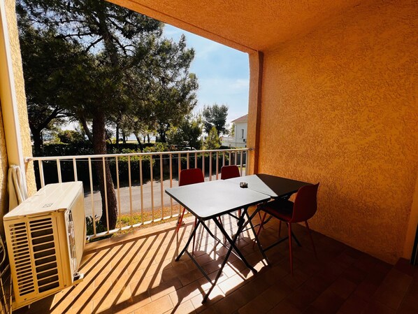 A balcony with a table and chairs, a white air conditioning unit, and a view of trees and a building. - Apartment, AC, sea view - 4 guests (Argelès-sur-Mer)
