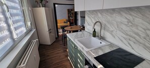 Une cuisine moderne dotée d’un évier encastré, un coin repas avec une table et des chaises, et une vue sur un salon avec un canapé et une bibliothèque, accessible par une porte ouverte.