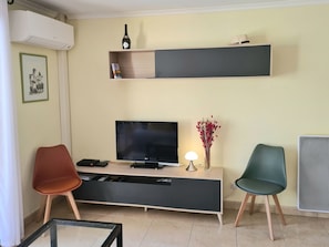 Ein Wohnzimmer mit Fernseher, einem Konsolentisch, zwei Sessel, einer Wandregal mit Flasche und Buch sowie einem Heizkörper.