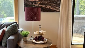 A cozy living room with a grey sofa, a round wooden coffee table, a red lamp, a decorative plate, and a painting of a sunset over a lighthouse.