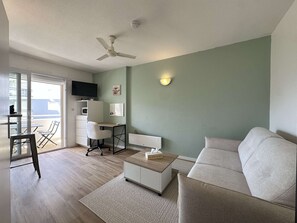 A living room with a sofa, a desk, a chair, a television, and a ceiling fan. - Studio in Minimes, parking - 2 guests (La Rochelle)
