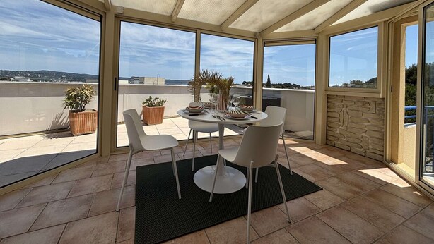 Un patio iluminado por el sol con una mesa para dos, ventanas amplias y plantas en macetas.