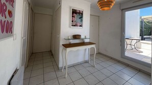 Un pasillo con una puerta blanca, un estante de cristal con una cesta, un cuadro enmarcado y una lámpara colgante.