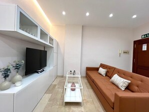 Ein modernes Wohnzimmer mit einem Flachbildfernseher, weißen Schränken, einem Glastisch und einem braunen Sofa mit gemusterten Kissen.