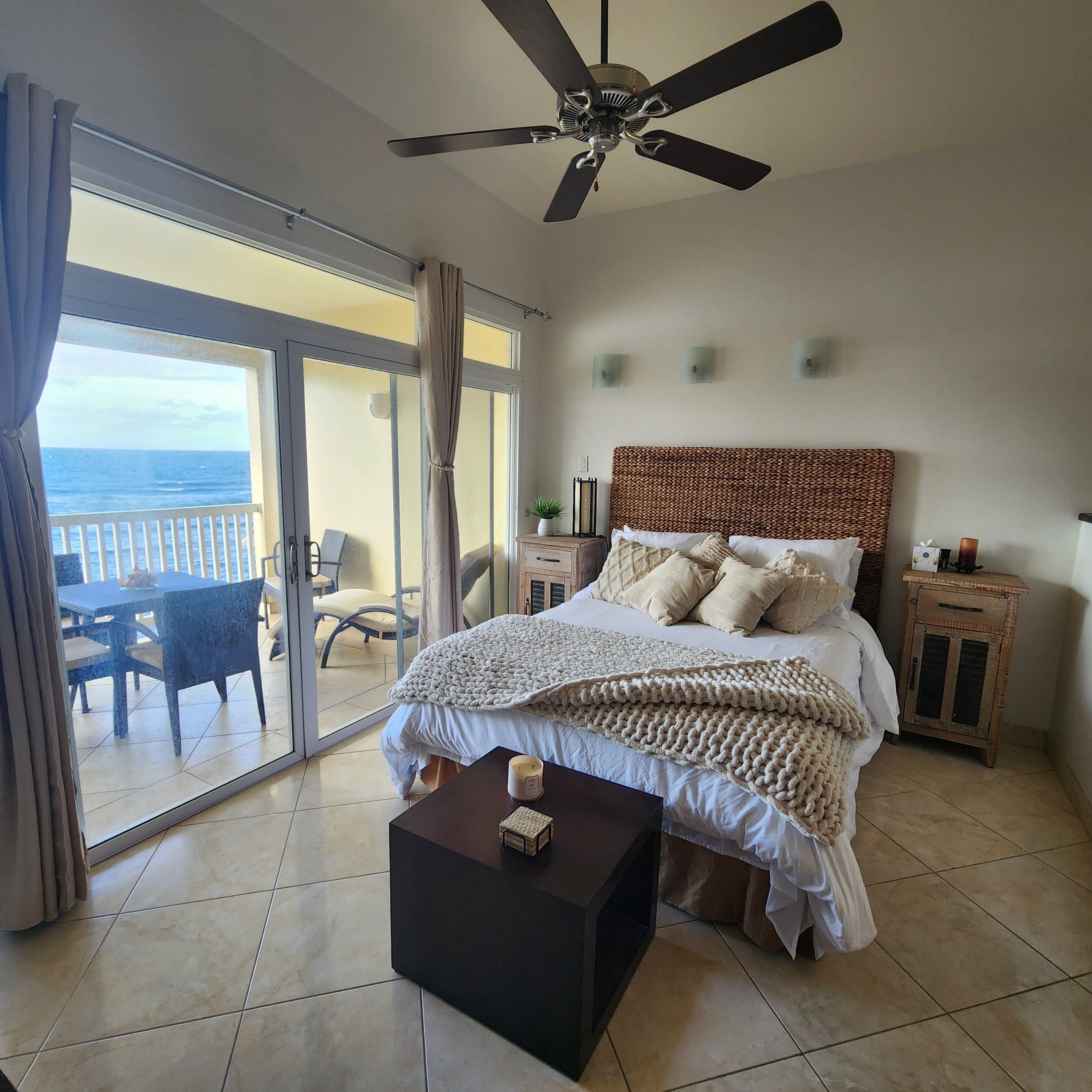 Une chambre avec un lit, un ventilateur de plafond, une table de chevet et une commode, et une vue sur l’océan par une porte-fenêtre coulissante.