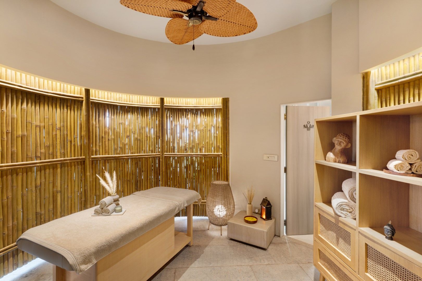 A spa room with a massage table, bamboo partitions, a ceiling fan, and shelves with towels and decorative items.