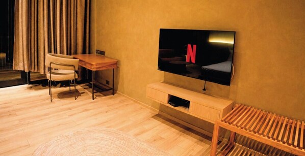 A modern living room with a flat-screen TV mounted on the wall, a wooden desk, a chair, and a small wooden bench.