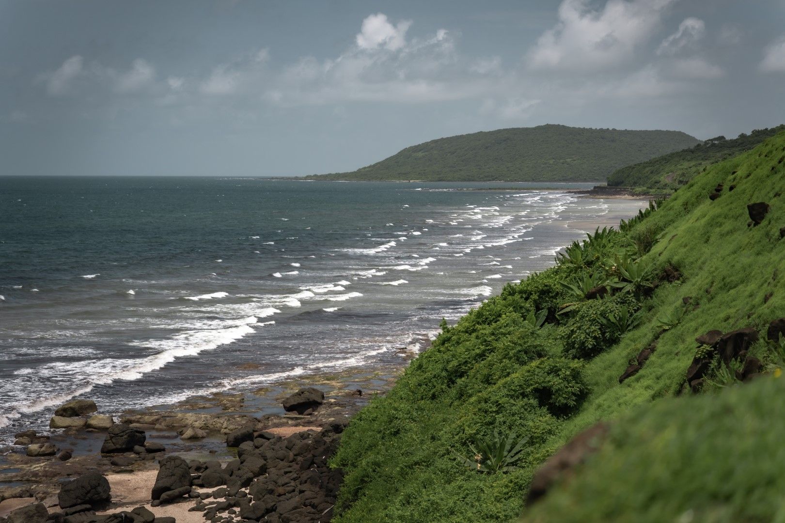 A coastal landscape with a rocky shoreline, waves, and a hill in the distance.