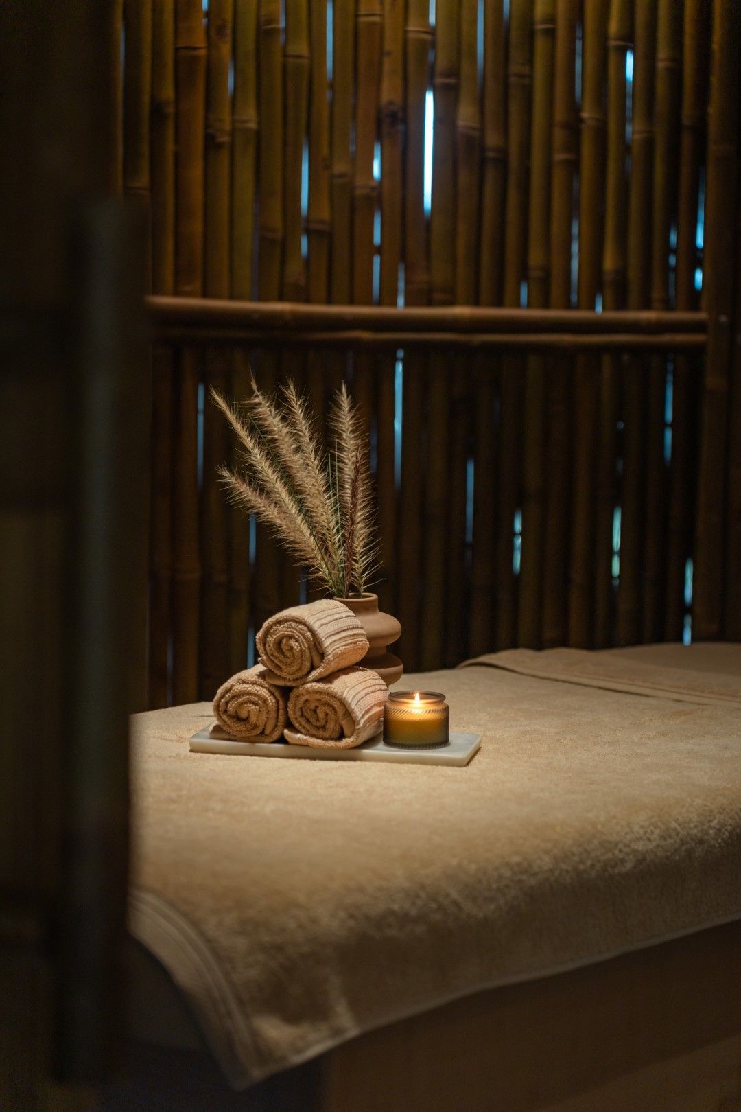 A spa setting with a massage table, rolled towels, a candle, and dried plants.