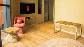 Una sala moderna con un televisor de pantalla plana, una butaca roja, un banco de madera y un tapete redondo tejido.