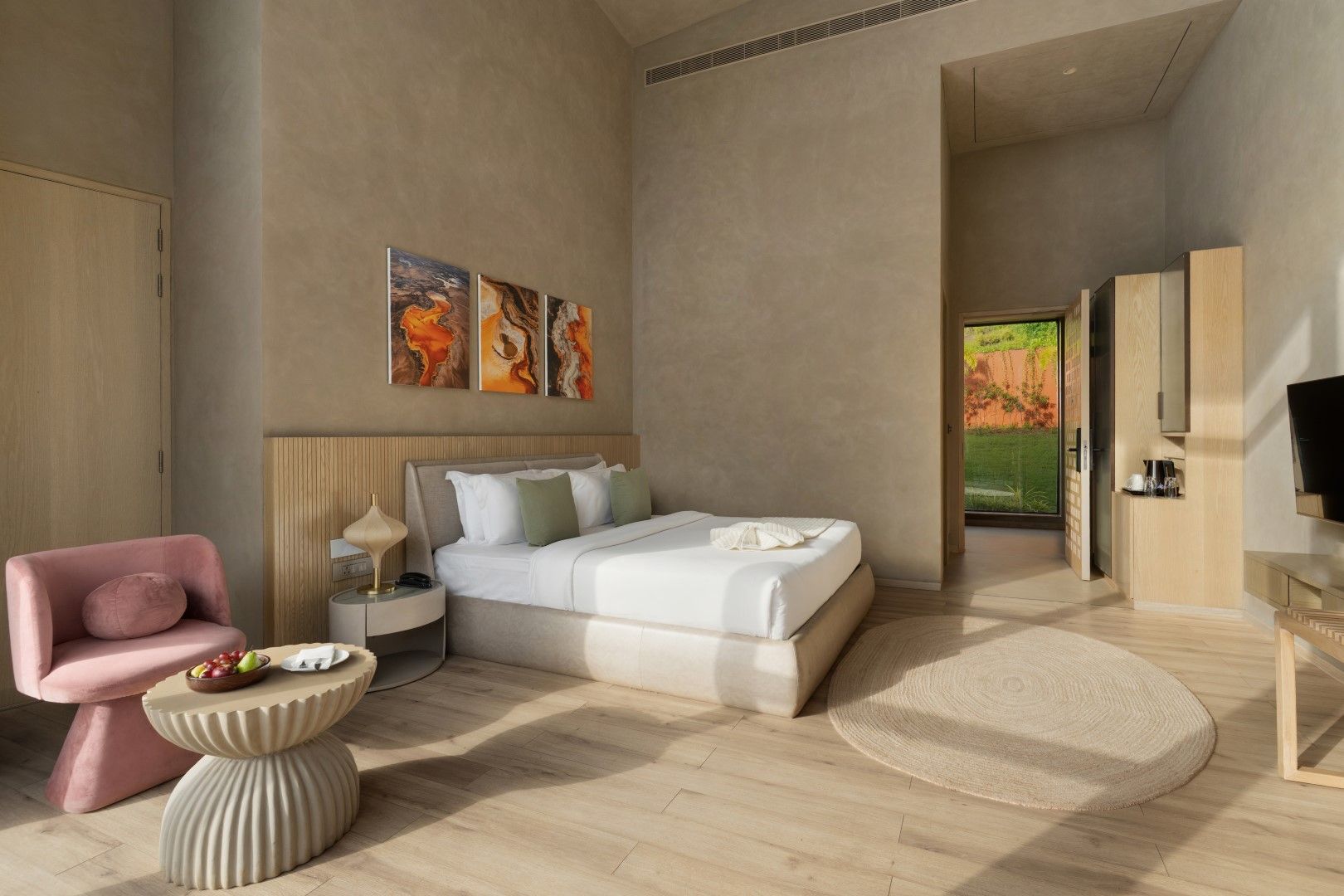 Une chambre d’hôtel moderne avec un grand lit, un fauteuil rose, une table de chevet et des œuvres d’art accrochées au mur.