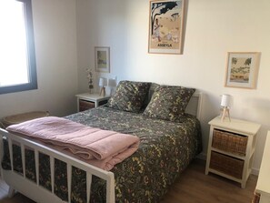 Ein Schlafzimmer mit einem Bett, einem Nachttisch, einer Lampe, einem gerahmten Bild und einem Fenster.