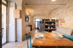 Un coin repas avec une table en bois, des chaises et un banc. On aperçoit, au fond, un espace cuisine avec des placards et un évier.