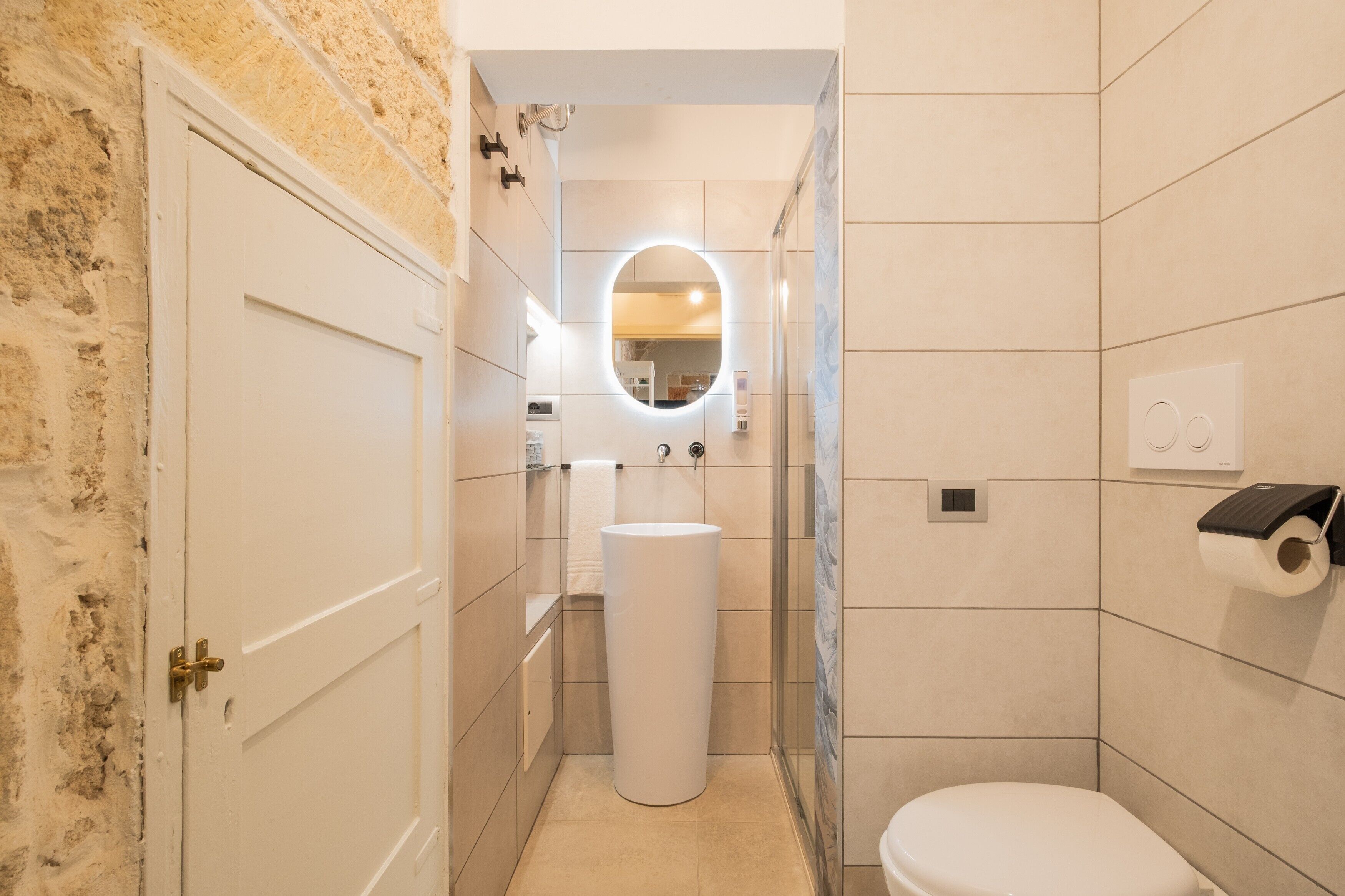 Un baño con lavamanos blanco, inodoro, espejo con luz y puerta.