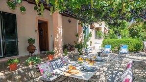 Patio esterno con tavolo apparecchiato per la colazione, circondato da piante in vaso e un'area salotto.