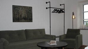 A room with a green sofa, a small round table, a coat rack, a floor lamp, and a painting on the wall.