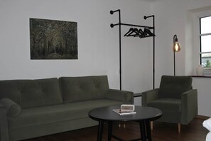 A room with a green sofa, a small round table, a coat rack, a floor lamp, and a painting on the wall.