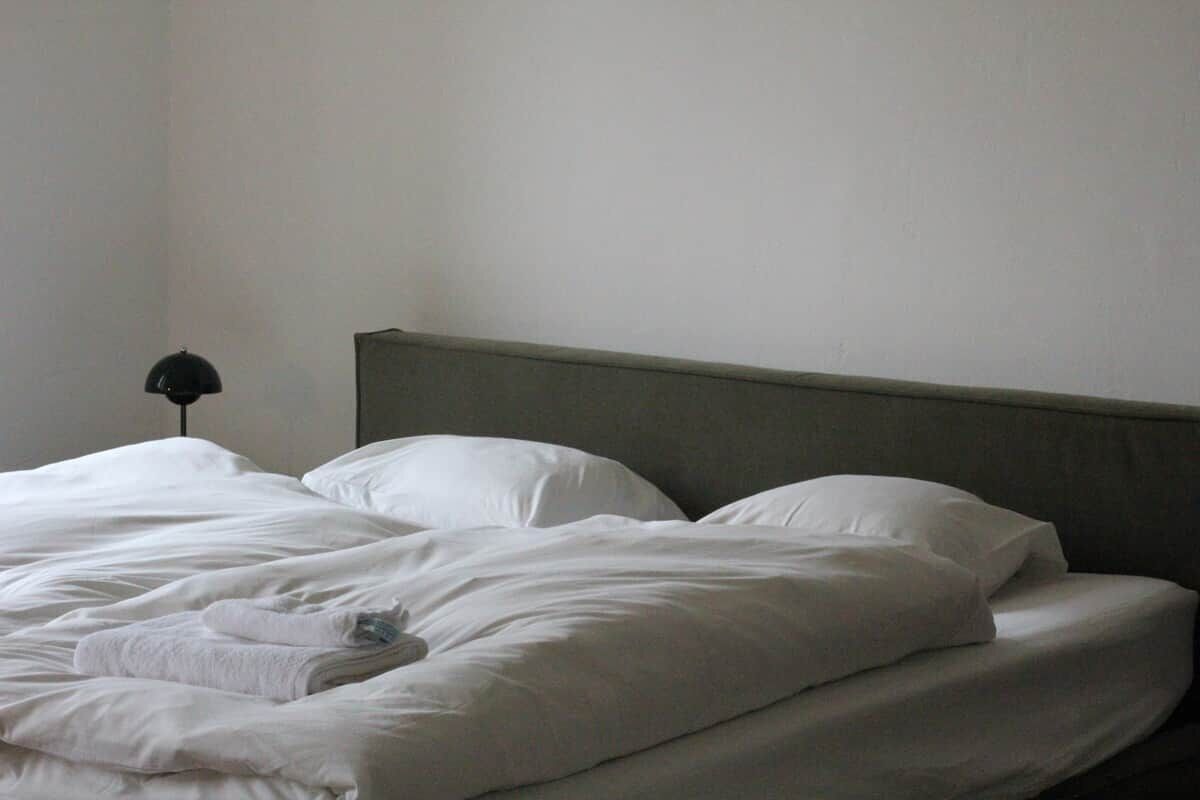 A bed with white linens and a grey headboard in a room with a plain wall and a single bedside lamp.