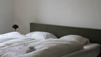 A bed with white linens and a grey headboard in a room with a plain wall and a single bedside lamp.