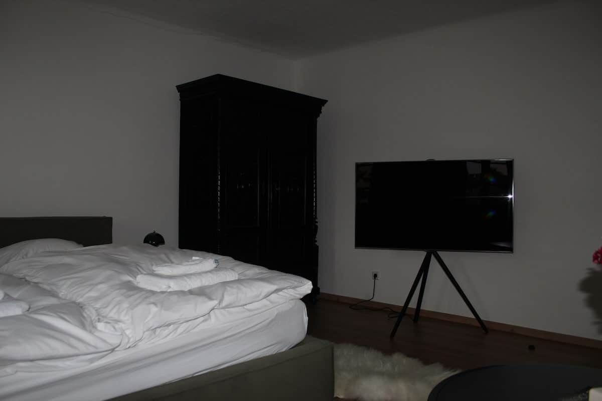 Une chambre à coucher avec un lit, une armoire sombre, un téléviseur posé sur un meuble et une petite table ronde.