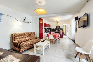Ein Wohnzimmer mit einer Couch, einem Couchtisch und einem Fernseher. Im Hintergrund ist eine Küchenzeile mit einem Esstisch und Stühlen zu sehen.