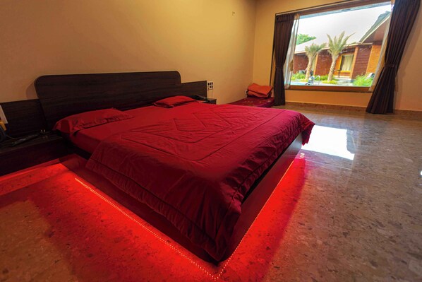 Un dormitorio con una cama grande, una mesita de noche, un ventanal con vistas a palmeras y una puerta.