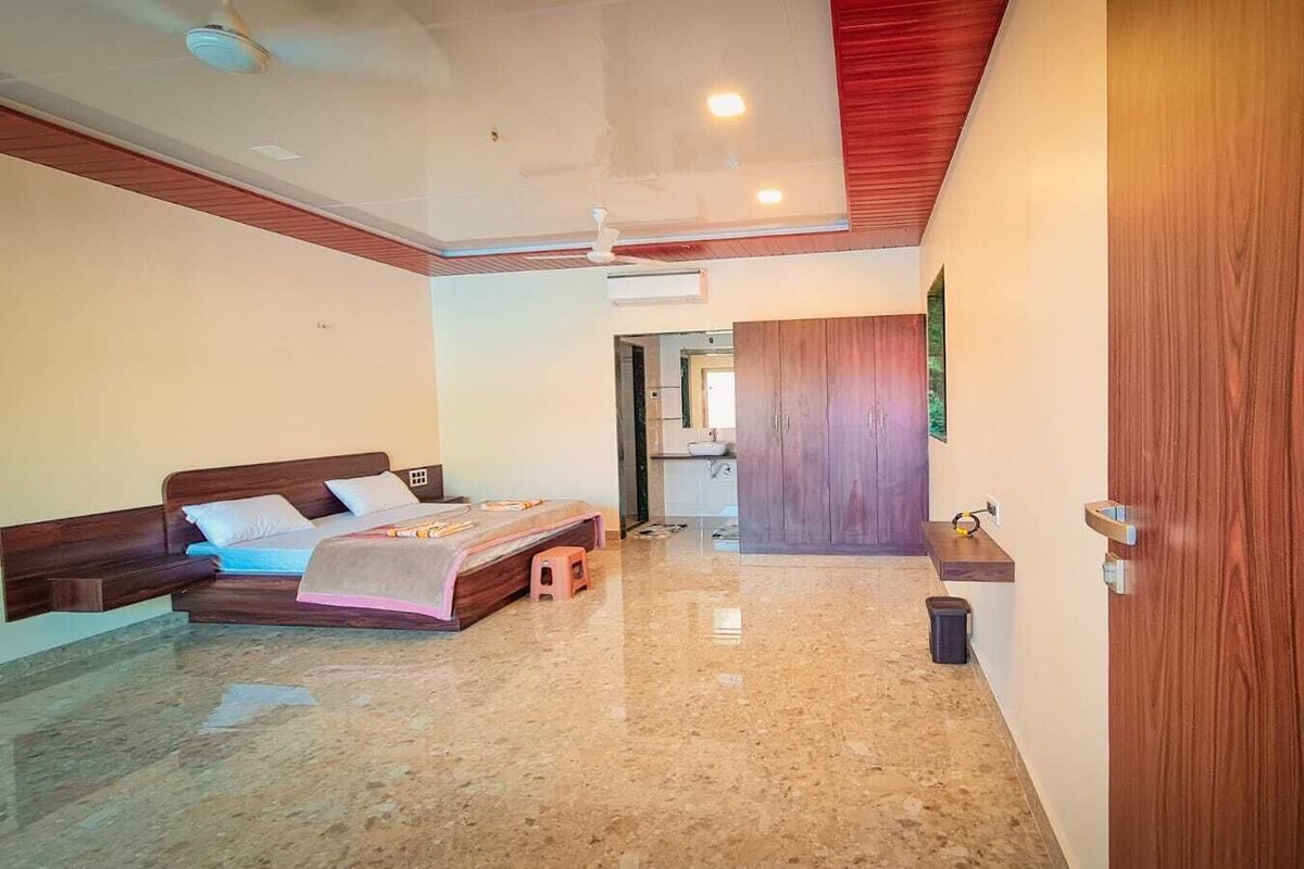 A bedroom with a bed, a small stool, a wooden door, and a hallway leading to a bathroom.
