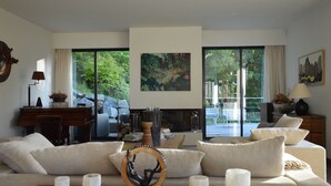 Una sala de estar moderna con sofá, mesa de centro y chimenea. Se observa un piano y un escritorio con lámpara. Amplias ventanas ofrecen vista a la vegetación exterior.