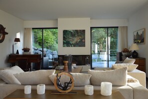 A modern living room with a sofa, coffee table, and fireplace. There is a piano and a desk with a lamp. Large windows offer a view of greenery outside. - Villa sea view Normandy Deauville (Trouville-sur-Mer)