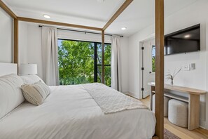 A modern bedroom with a large bed, a TV, and a view of trees through the window. - Iconic Nashville Twin Townhomes | 6BR + Hot Tubs (Nashville)