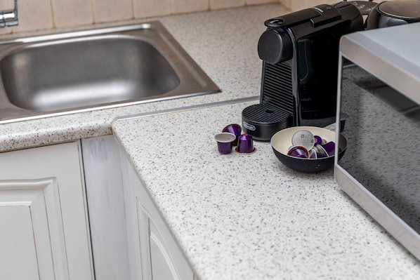 A kitchen with a stainless steel sink, a coffee maker, and a bowl with cups on a speckled countertop. - Mary & Jo's Apartments (Naxos)